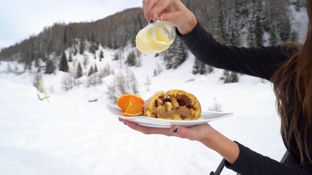 Beautiful girl pouring vanilla sauce over strudel cake on snowy mountains. In slow motion
