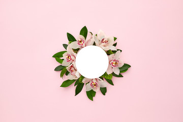 White round frame decorated with green leaves and orchid flowers on pink pastel background. empty...