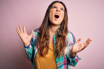 Young beautiful brunette woman wearing casual colorful shirt standing over pink background crazy and mad shouting and yelling with aggressive expression and arms raised. Frustration concept.