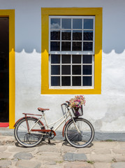 Fototapeta na wymiar Beautiful bicycle with flowers parked in front of a yellow window in the historic city of Paraty, Brazil.