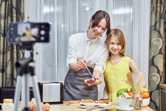 Woman with her child vlogging in kitchen, mother smiles daughter, look at camera, cooking tosts with jam.