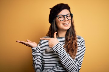 Young beautiful brunette woman wearing french beret and glasses over yellow background amazed and smiling to the camera while presenting with hand and pointing with finger.