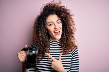 Young beautiful woman with curly hair and piercing doing coffee holding french coffeemaker very happy pointing with hand and finger