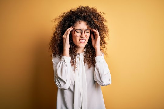 Young beautiful brunette woman with curly hair and piercing wearing shirt and glasses with hand on head for pain in head because stress. Suffering migraine.