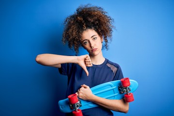 Young sporty woman with curly hair and piercing holding skate over blue background with angry face,...