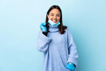 Young brunette mixed race woman protecting from the coronavirus with a mask and gloves over isolated blue background laughing