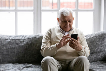 Smiling positive old man sit on couch in living room holding mobile phone enjoy usage feels...