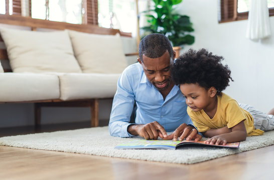 Happy African American father and son are reading a book and smiling while lying on floor spending time together at home. Children education and development concept.