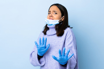 Young brunette mixed race woman protecting from the coronavirus with a mask and gloves over isolated blue background nervous stretching hands to the front