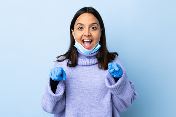 Young brunette mixed race woman protecting from the coronavirus with a mask and gloves over isolated blue background surprised and pointing front