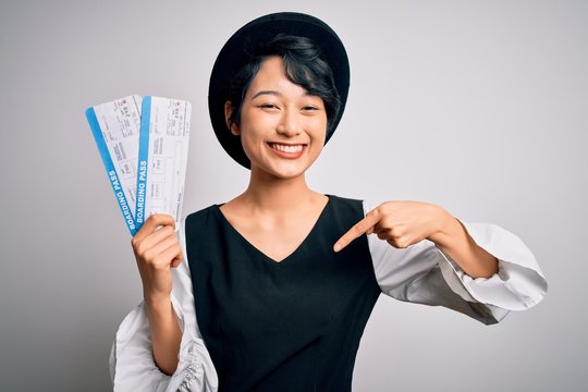 Young beautiful chinese tourist woman holding boarding pass airlane over white background with surprise face pointing finger to himself