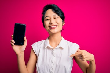 Young beautiful asian girl holding smartphone showing screen over isolated pink background with surprise face pointing finger to himself