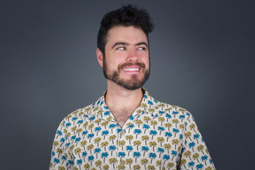 Dreamy young male with thoughtful expression, looks away keeps hands down bitting his lip thinks about something pleasant poses against gray wall.