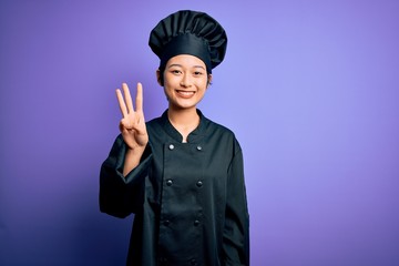 Young beautiful chinese chef woman wearing cooker uniform and hat over purple background showing and pointing up with fingers number three while smiling confident and happy.