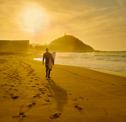 A surfer walking on the Zurriola Beach at sunset with the Monte Urgull in the background. San...