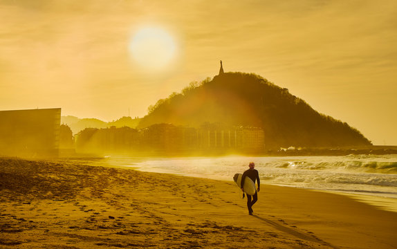 A surfer walking on the Zurriola Beach at sunset with the Monte Urgull in the background. San Sebastian, Basque Country, Guipuzcoa. Spain.