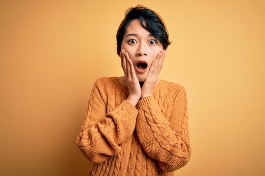 Young beautiful asian girl wearing casual sweater and diadem standing over yellow background afraid and shocked, surprise and amazed expression with hands on face