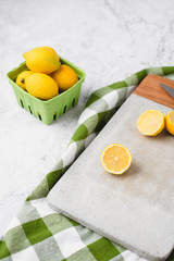 Fresh Lemons in a Green Produce Basket on a White Marble Countertop; Some Cut Open on a Cement Cutting Board; Green and White Checked Kitchen Towel in Background