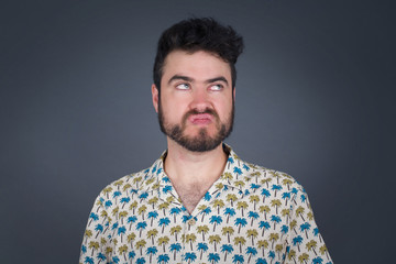 Gloomy, wondering young man face looking up lips together, being upset thinking about something important, keeps hands down, man standing indoors.