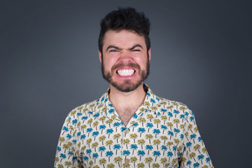 Fototapeta premium Mad crazy man clenches teeth angrily, being annoyed with coming noise, dressed in fashionable clothes, isolated over gray background. Negative feeling concept.