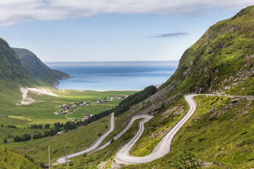 Mountain road with many curves leading to a white sand beach on the banks of a deep blue fjord, on a beautiful summer day in Norway with clear blue skies, abundant green vegetation