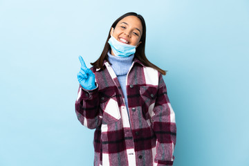 Young brunette mixed race woman protecting from the coronavirus with a mask and gloves over isolated blue background showing and lifting a finger in sign of the best