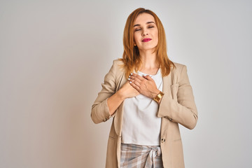 Redhead caucasian business woman standing over isolated background smiling with hands on chest with closed eyes and grateful gesture on face. Health concept.