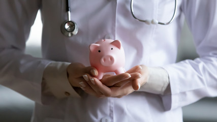 Hands of therapist in white coat uniform holding piggy bank close up horizontal cropped image, concept of paid medicine services, free medical care, save money special offer health check-up in clinic - obrazy, fototapety, plakaty