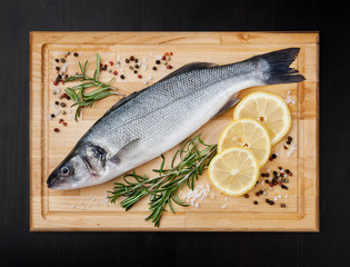 Fresh uncooked seabass with lemon, rosemary and spice on wooden board over black backdground