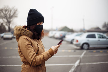 Young girl in city street wearing black sterile medical face mask. Woman using the phone to search for news about nCov 2019. Quarantine COVID-19 pandemic coronavirus epidemic and health care concept.