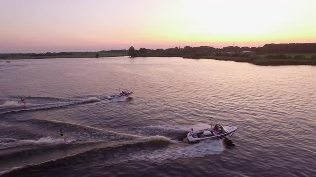Two speed boats with beginner waterskiiers and wakeboarder behind it during beautiful sunset