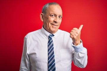 Senior handsome businessman wearing elegant tie standing over isolated red background smiling with happy face looking and pointing to the side with thumb up.