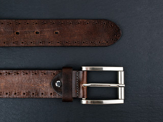 End parts of brown genuine leather belt with classic metal buckle on a black background. Men's stylish leather accessories. Low key image.
