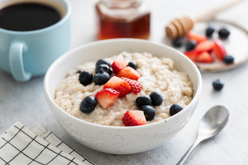 Oatmeal porridge with berries, honey and cup of coffee on white table. Healthy breakfast food,...