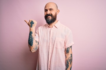 Handsome bald man with beard and tattoo wearing casual shirt over isolated pink background smiling with happy face looking and pointing to the side with thumb up.