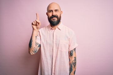 Handsome bald man with beard and tattoo wearing casual shirt over isolated pink background showing and pointing up with finger number one while smiling confident and happy.