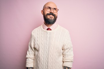 Handsome bald man with beard and tattoo wearing glasses and sweater over pink background looking away to side with smile on face, natural expression. Laughing confident.