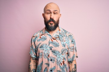 Obraz premium Handsome bald man with beard and tattoo wearing casual floral shirt over pink background making fish face with lips, crazy and comical gesture. Funny expression.