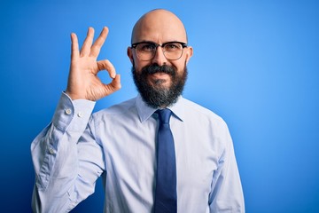Handsome business bald man with beard wearing elegant tie and glasses over blue background smiling positive doing ok sign with hand and fingers. Successful expression.