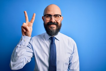 Handsome business bald man with beard wearing elegant tie and glasses over blue background showing and pointing up with fingers number two while smiling confident and happy.