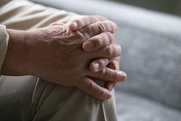 Fototapeta premium Old man sitting on couch close up view palms folded on knee. Concept of older generation lonely person, senile diseases osteoarthritis rheumatoid arthritis, male during psychological therapy session