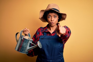 Young African American farmer woman with curly hair wearing apron and hat using water can pointing with finger to the camera and to you, hand sign, positive and confident gesture from the front