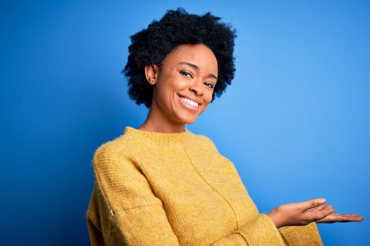 Young beautiful African American afro woman with curly hair wearing yellow casual sweater Inviting to enter smiling natural with open hand