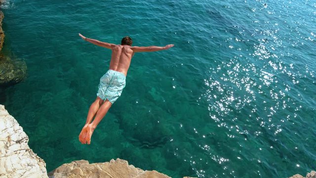 SLOW MOTION, COPY SPACE: Young man on a relaxing summer vacation in Croatia does cliff diving on a sunny day. Cinematic shot of a man jumping off a rocky ledge and into the glistening blue ocean.