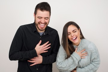 Joyful European student keeps hands crossed, laughs at good joke, wears casual clothes and round spectacles, standing indoors. Happy young woman with long hair poses against gray wall.