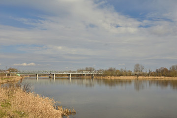 Brzeg rzeki porośnięty lasem w słoneczny dzień z chmurami na niebie, w tle most przez rzekę.