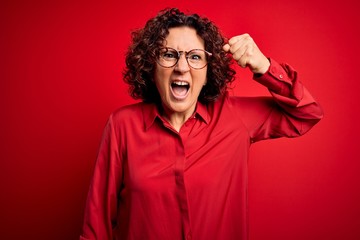 Fototapeta na wymiar Middle age beautiful curly hair woman wearing casual shirt and glasses over red background angry and mad raising fist frustrated and furious while shouting with anger. Rage and aggressive concept.