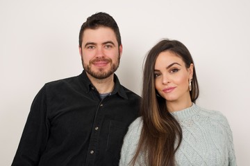 Close up studio shot of beautiful young couple looking at camera with charming cute smile while posing against white blank copy space wall for your content