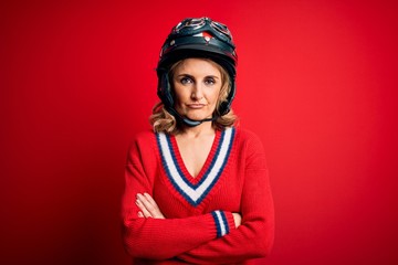 Middle age beautiful blonde motorcyclist woman wearing moto helmet over red background skeptic and...