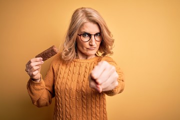 Middle age beautiful blonde woman eating energetic protein bar over yellow background annoyed and...